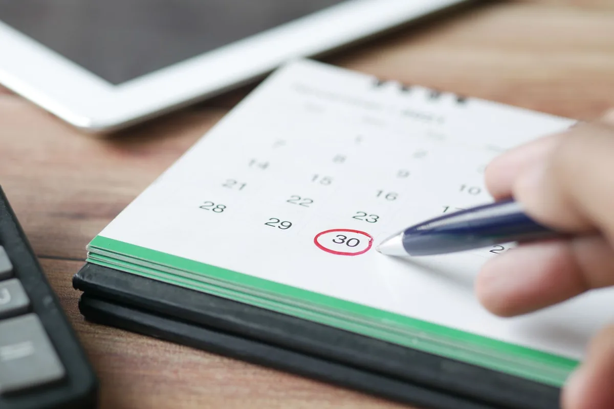 Une main tient un stylo près d'un calendrier de bureau dont une date est entourée en rouge, posé sur une table en bois à côté d'une tablette.