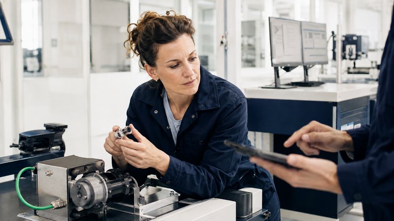 Technicienne en atelier industriel examinant une pièce mécanique, un collègue lui présentant une tablette de contrôle