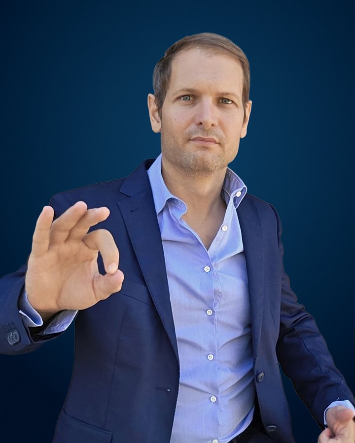 Benjamin Duplaa en veste bleu marine et chemise bleu clair au col ouvert, regard face à l'objectif, une main levée, pouce et index presque joints, sur un fond bleu nuit